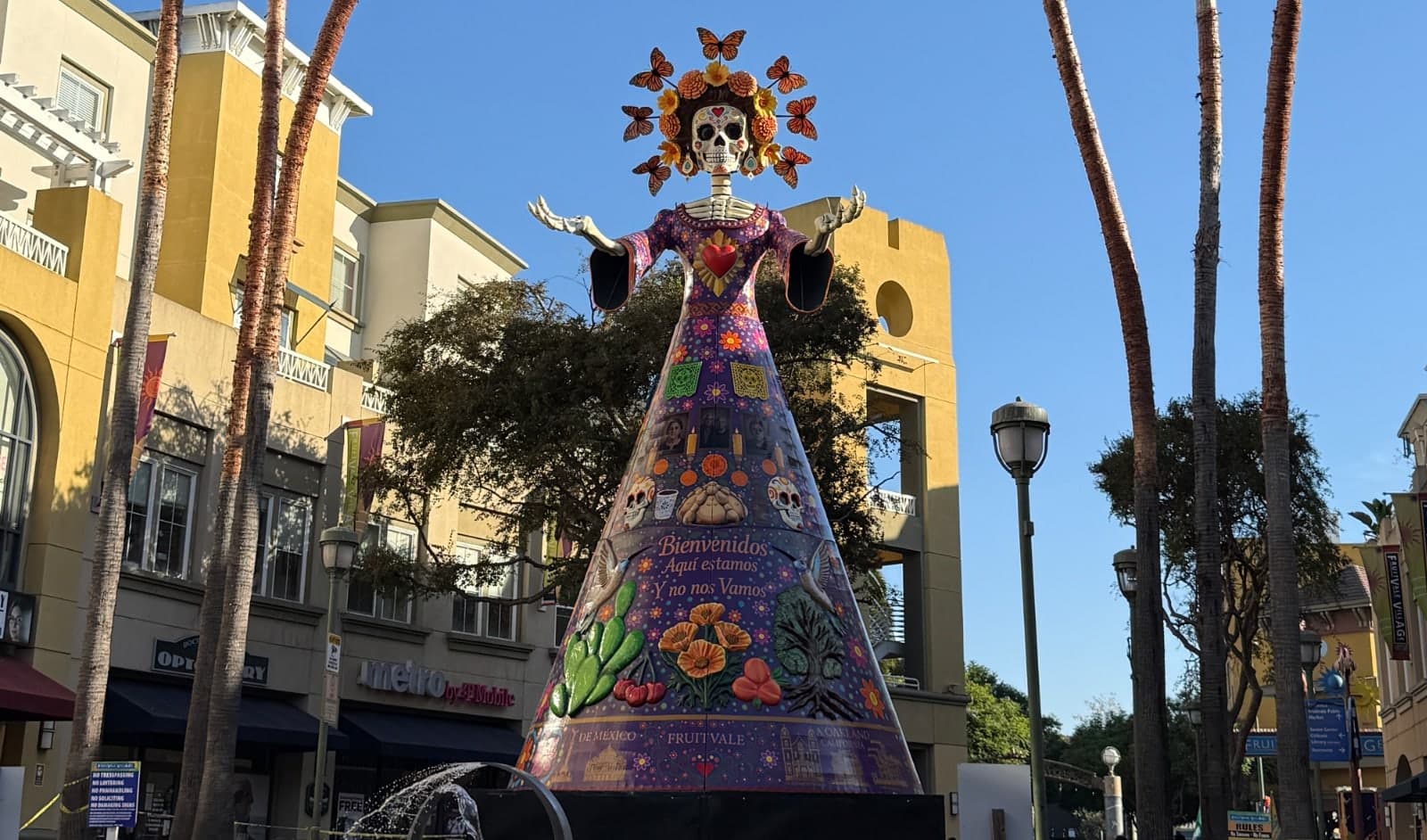 Giant La Catrina statue with a colorful floral dress and butterfly headdress, surrounded by palm trees and buildings in Fruitvale, Oakland.